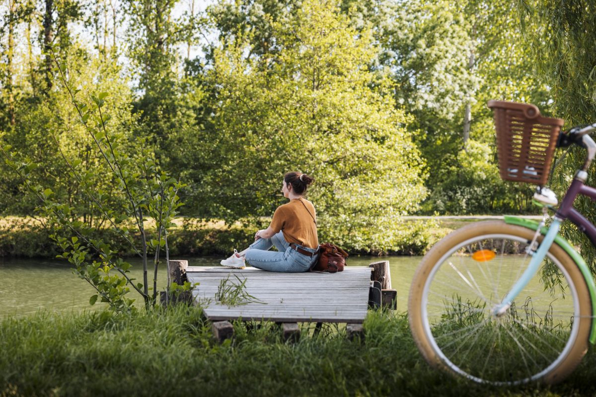 Jeune fille assise sur un ponton au bord d'un canal du Marais poitevin, son vélo, tout proche, est posé contre un arbre.